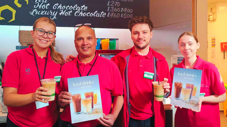 4 National Trust staff members, standing with Iced Coffees and signs that state, 'Iced drinks are back' within the Cafe area of the Giant's Causeway Visitor Centre